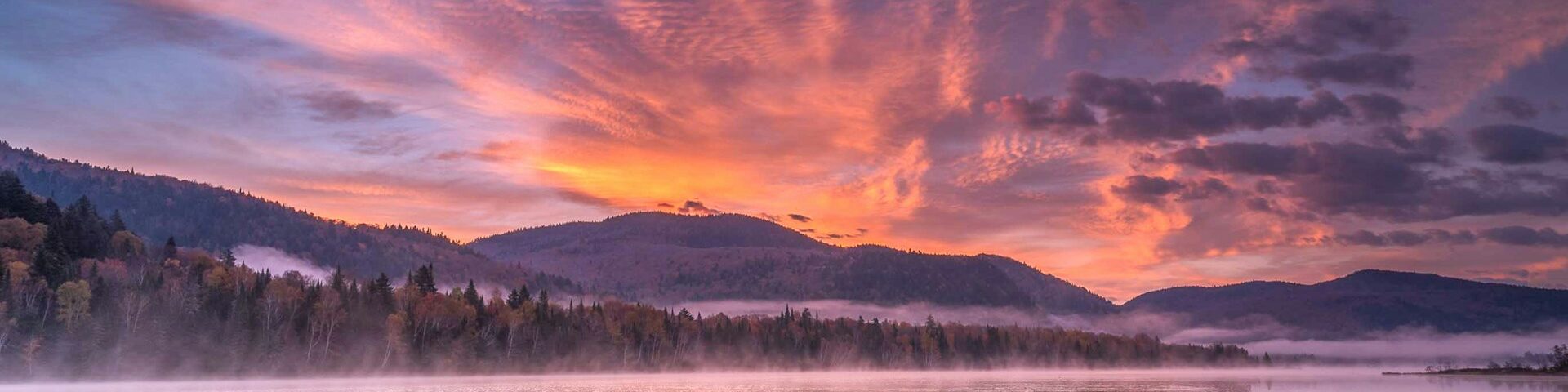 Un lac en hiver avec une montagne au loin pendant le levé du soleil. Les nuages sont oranges et une brumes flotte au-dessus du lac et de la forêt